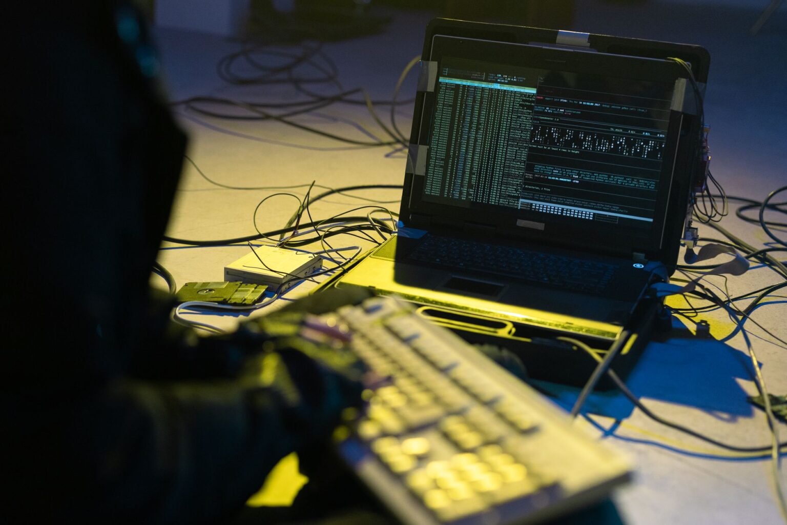 Haker siedzący przed laptopem z widocznym kodem i kablami na podłodze w ramach zagrożenia cyberbezpieczeństwa
